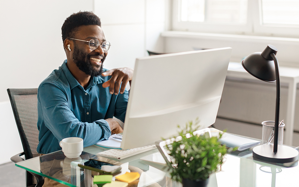 Handsome black man entrepreneur sitting at desk in office, looking at computer monitor and smiling, having video call